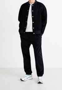 Svart bomberjacka med silverknappar, vit texturerad tröja, svarta byxor och vita sneakers med gröna detaljer.
