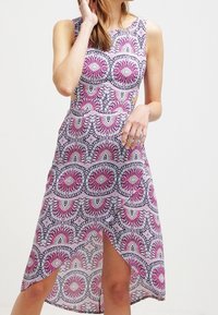 Kvinna i en ärmlös klänning med rosa, lila och svarta geometriska solstrålemönster och framförslits, stående mot en enhetlig bakgrund.