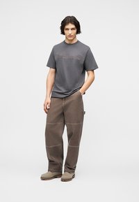 Νεαρός άνδρας που στέκεται με ένα χέρι στην τσέπη, φοράει γκρι μπλουζάκι Timberland, καφέ φαρδιά παντελόνια και μπεζ μποτάκια, πάνω σε λευκό φόντο.