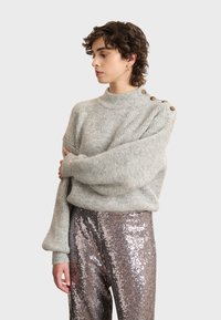 Svijetlosivi pleteni pulover s visokim ovratnikom i oversized rukavima, s tri smeđa dugmeta na ramenu. Uparen s hlačama od šljokica.