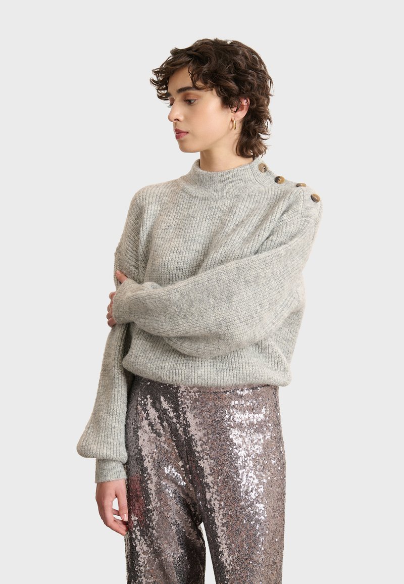 Hellgrauer, gestrickter Pullover mit hohem Kragen und übergroßen Ärmeln, der drei braune Knöpfe an der Schulter hat. Kombiniert mit Pailletten-Hosen.