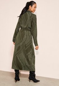 Rochie lungă de culoare verde măslinie, cu un model ondulat alb, mâneci lungi și talie evidențiată cu un brâu. Purtați cu cizme negre cu toc. Material texturat.