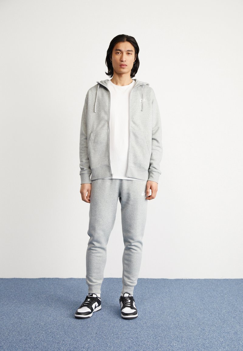 Hoodie zippé gris avec cordons de serrage, porté sur un t-shirt blanc et un jogging assorti. Des baskets noires et blanches complètent la tenue.