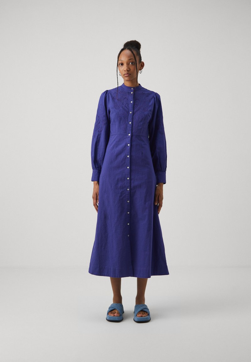 Abito button-up a maniche lunghe in lino blu scuro, con dettagli ricamati e una silhouette fluida. Abbinato a sandali blu a scivolo.