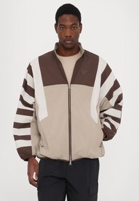 Béžová a hnědá bunda s bílými akcenty, přední zip, stojací límec a široké pruhované rukávy, vyrobená z lehké látky.