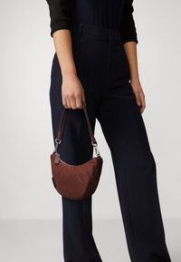 Bruine nylon handtas met een gebogen vorm, korte bruine leren band en zilverkleurige metalen details, vastgehouden door een persoon die donkere broek draagt.