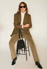 Blazer marron porté sur un chemisier blanc à volants, jean olive, sac seau noir à franges, bottines noires et lunettes de soleil foncées.