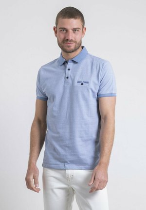 Homme aux cheveux bruns courts portant un polo bleu clair à motifs et un pantalon blanc, debout devant un fond clair uni.