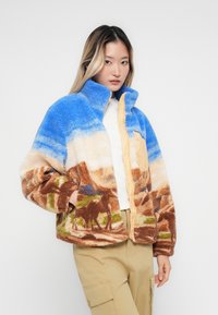 Jeune femme portant une veste polaire colorée avec un motif de paysage bleu, blanc et marron et un pantalon cargo beige, posant les mains dans les poches.