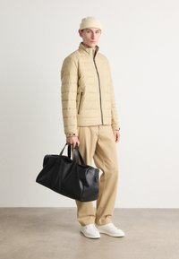 Jachetă puffer bej cu cusături orizontale, fermoar negru, pantaloni bej, adidași albi și o geantă mare de voiaj din piele neagră.