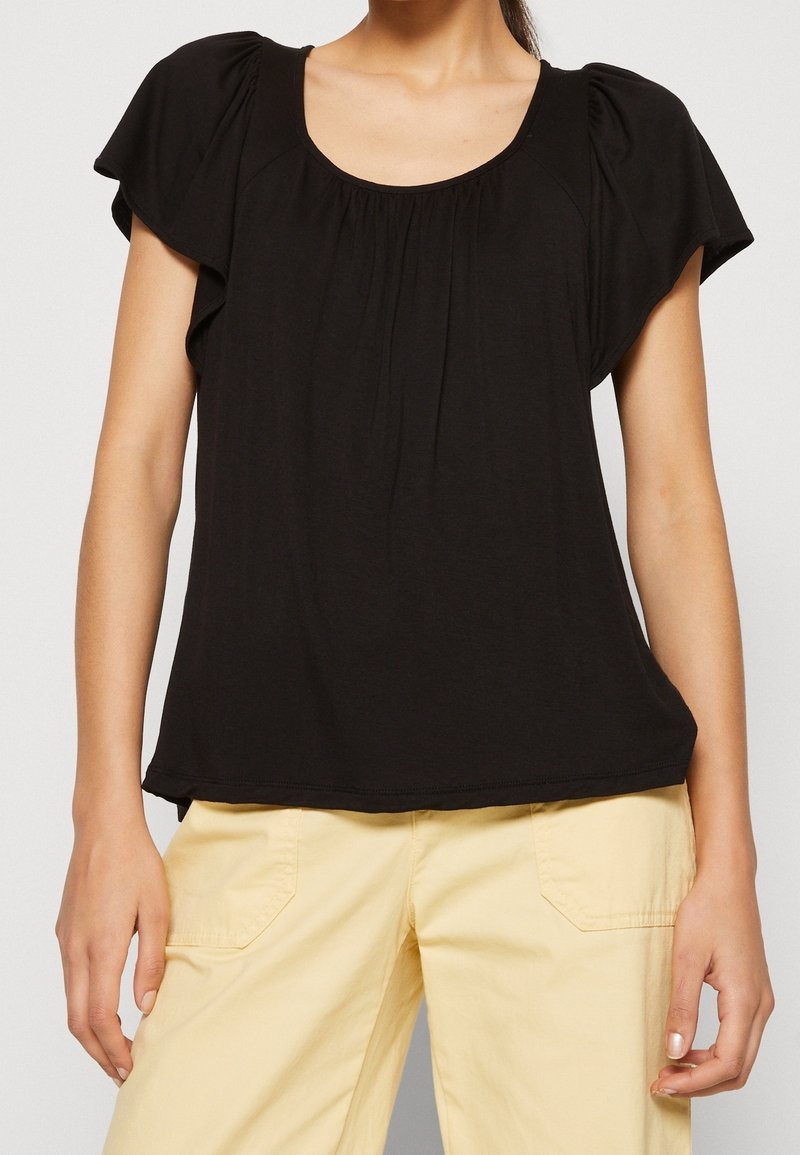 Femme portant un haut noir ample à manches courtes avec des fronces subtiles et un pantalon beige clair avec des poches avant.