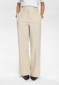 Pantaloni larghi beige con vita alta, tasche laterali, tessuto morbido e dettaglio della piega; indossati con scarpe nere a punta.