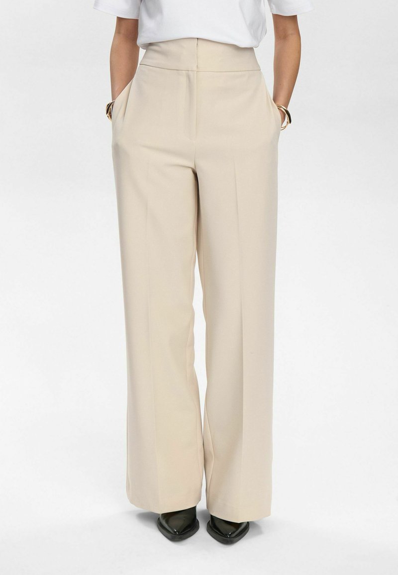 Pantaloni larghi beige con vita alta, tasche laterali, tessuto morbido e dettaglio della piega; indossati con scarpe nere a punta.