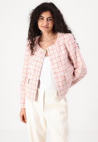 Rosa og hvit rutet tweedjakke med gullknapper, rund hals, og sidelommer, kombinert med en hvit blondebluse og stripete bukser.