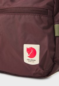 Zaino bordeaux con una superficie in tessuto testurizzato, caratterizzato da un patch del logo Fjällräven in crema, una silhouette di volpe rossa e dettagli in verde sulle cinghie.