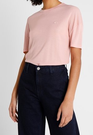 Vrouw die een lichtroze T-shirt met korte mouwen draagt, ingestopt in een donkerblauwe high-waisted spijkerbroek tegen een effen achtergrond.