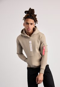 Beige hettegenser med snor, frontlomme og lommetil flap på ermet; har "ALPHA" merke og rød tag; glatt bomullsblanding stoff.