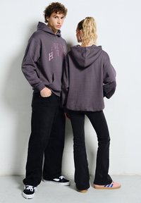 Młody mężczyzna i kobieta ubrani w ciemne bluzy z kapturem oraz czarne spodnie, stojący na tle białej ściany, w casualowych trampkach.