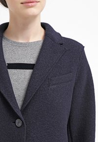 Laivastonsininen villainen bleiseri teksturoidulla pinnalla, yksinappinen suljenta ja notkoviistot kaulukset. Siinä on pieni rintatasku ja näkyvä nappidetalji.