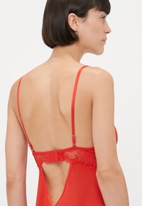 Femme aux cheveux courts et foncés portant de la lingerie en dentelle rouge à fines bretelles ajustables, dévoilant les détails du dos et de l'épaule sur fond blanc.