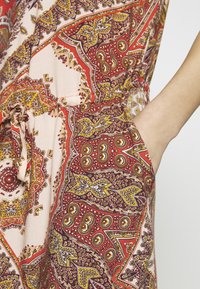 Mano que lleva un vestido con un bolsillo estampado que presenta diseños de paisley en rojo, amarillo y morado, con un detalle de cintura atada.
