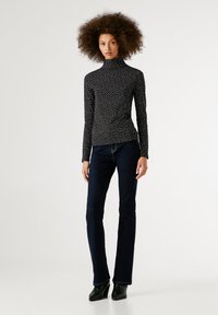 Göndör hajú nő fekete-fehér pöttyös garbót, sötétkék farmernadrágot és fekete magassarkú csizmát visel, háttérben egy fehér fal áll.