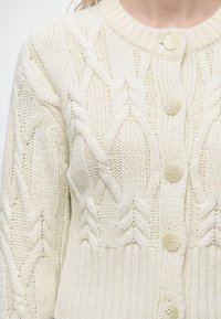 Gros plan d’un cardigan écru en maille torsadée avec de gros boutons et des motifs tressés texturés sur le devant et la manche.