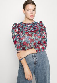 Blusa floreale con maniche a sbuffo in motivi blu, rossi e verdi. Collo arruffato, abbinata a jeans in denim blu a vita alta.