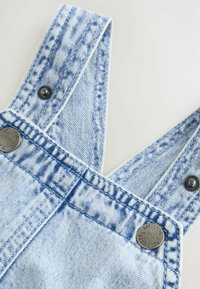 Ljusblå denimsoveralls med justerbara axelremmar, synlig sömnad och silverknappar med ingraverad text. Strukturerad tyg med blekt utseende.