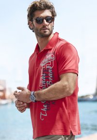 Rotes Poloshirt mit aufgedruckten Grafiken, kurzen Ärmeln und Seitenschlitzen. Trägt eine Sonnenbrille und eine gestreifte Uhr. Im Hintergrund ist Wasser zu sehen.