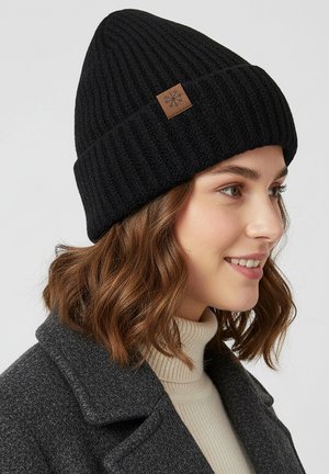 Black knitted beanie with ribbed texture, featuring a brown leather patch. Worn by a person with shoulder-length brown hair.