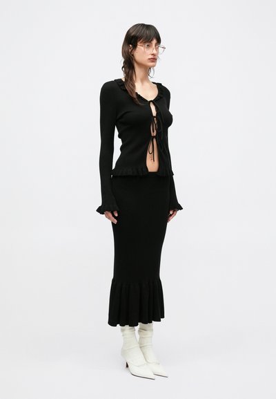 Mustast soonikkoeelsest kampsunist, millel on kroogitud kaelus ja ees paelaga kinnituv detail. Sobiva musta seelikuga, millel on lehviv serv. Modell kannab valgeid kontsakingi ja kreemjaid sokke.