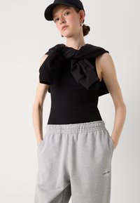 Zwarte geribbelde tanktop met grijze sportbroek met elastische tailleband en zijzakken. Zwarte sweatshirt over de schouders gedrapeerd. Zwarte pet.
