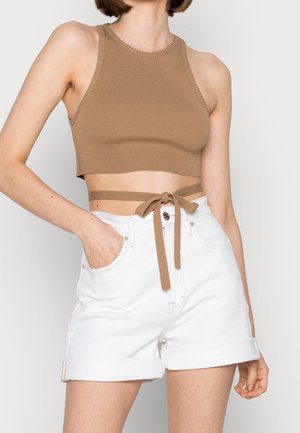Mujer con un top corto marrón sin mangas con detalle anudado en la cintura y shorts blancos de tiro alto con dobladillo, posando con una mano en el bolsillo.