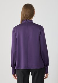 Bluză din satin violet cu mâneci lungi, gât înalt și manșete cu nasturi; textură netedă și un luciu subtil. Detaliile cusăturii din spate sunt vizibile.