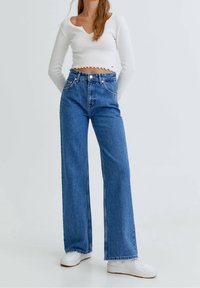 Top corto blanco de manga larga con dobladillo en forma de concha, combinado con unos vaqueros de pierna ancha azules. Los vaqueros tienen una cintura alta y una textura clásica de denim. Zapatillas blancas.