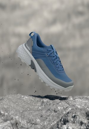 Modrá a sivá turistická obuv Columbia s hrubou podrážkou zavesená nad kamenistým terénom, pričom okolo lietajú častice špiny.