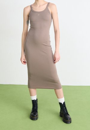 Personne portant une robe midi taupe ajustée sans manches, des chaussettes blanches et des bottines noires à lacets sur un sol vert.