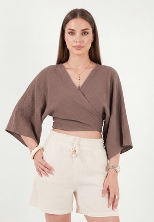 Femme aux longs cheveux portant un haut cache-cœur marron à larges manches et un short blanc avec cordon, debout avec une main sur la hanche.