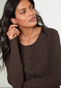 Haut à manches longues brun foncé côtelé avec un col arrondi et cinq boutons noirs sur le devant. Texture de tissu douce.