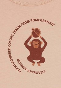 Helles rosa Stoff mit einem braunen Affen-Logo, der einen Granatapfel hält, umgeben von dem Text "Pflanzenbasierte Farben vom Granatapfel!"