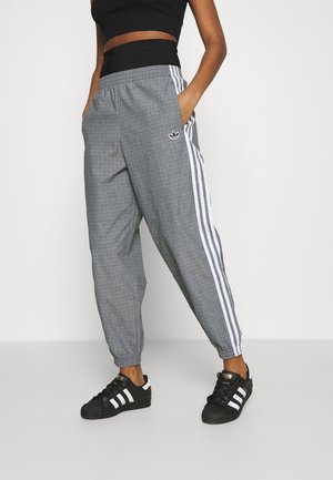 Person trägt graue Adidas-Trainingshose mit weißen Streifen, schwarzes Crop-Top und schwarze Adidas-Sneakers, steht vor weißem Hintergrund.