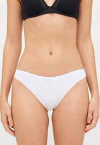Culottes de bikini en coton blanc avec dentelle en bord scallopé, présentant une ceinture douce embossée du nom de la marque. Texture lisse, coupe classique.