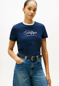 Marineblå bomulds T-shirt med hvid rund halsudskæring og "Hilfiger" logo. Kombinationen er med høj talje, lys blå denim jeans og sort bælte.