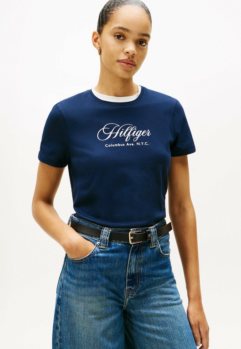 Marineblå bomulds T-shirt med hvid rund halsudskæring og "Hilfiger" logo. Kombinationen er med høj talje, lys blå denim jeans og sort bælte.