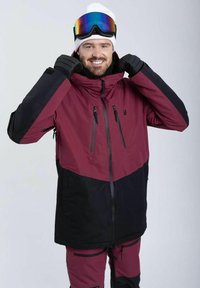 Mørkerød og sort skijakke med høj krave, lynlåse på brystet og hætte. Model bærer skibriller og handsker.