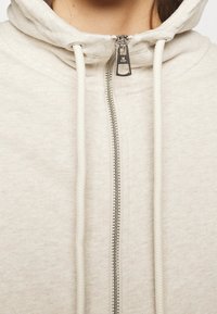 Hoodie beige chiaro realizzato in tessuto morbido, con zip frontale completa, cappuccio regolabile con lacci e finitura texture. Accentuato da hardware argentato.