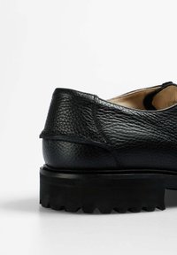 Zapato de cuero negro con parte superior texturizada, forma redondeada y suela de goma gruesa y estriada. Vista lateral que resalta la costura y el detalle del tacón.