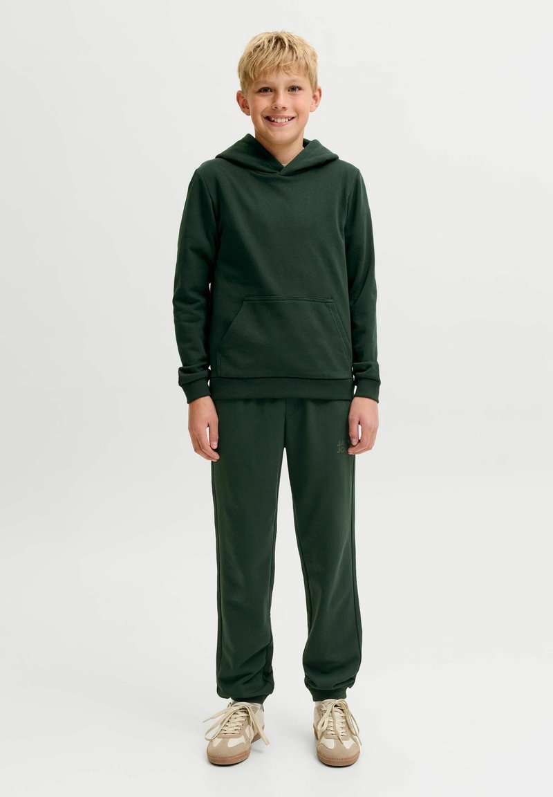 Garçon blond souriant, portant un sweat à capuche vert foncé et un pantalon de survêtement assorti avec des baskets beiges, debout sur un fond clair uni.