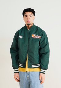 Grøn bomberjakke med broderet "Manhattan Ducks" logo, stribede manchetter, knaplukning, båret over en gul skjorte og lyse jeans.
